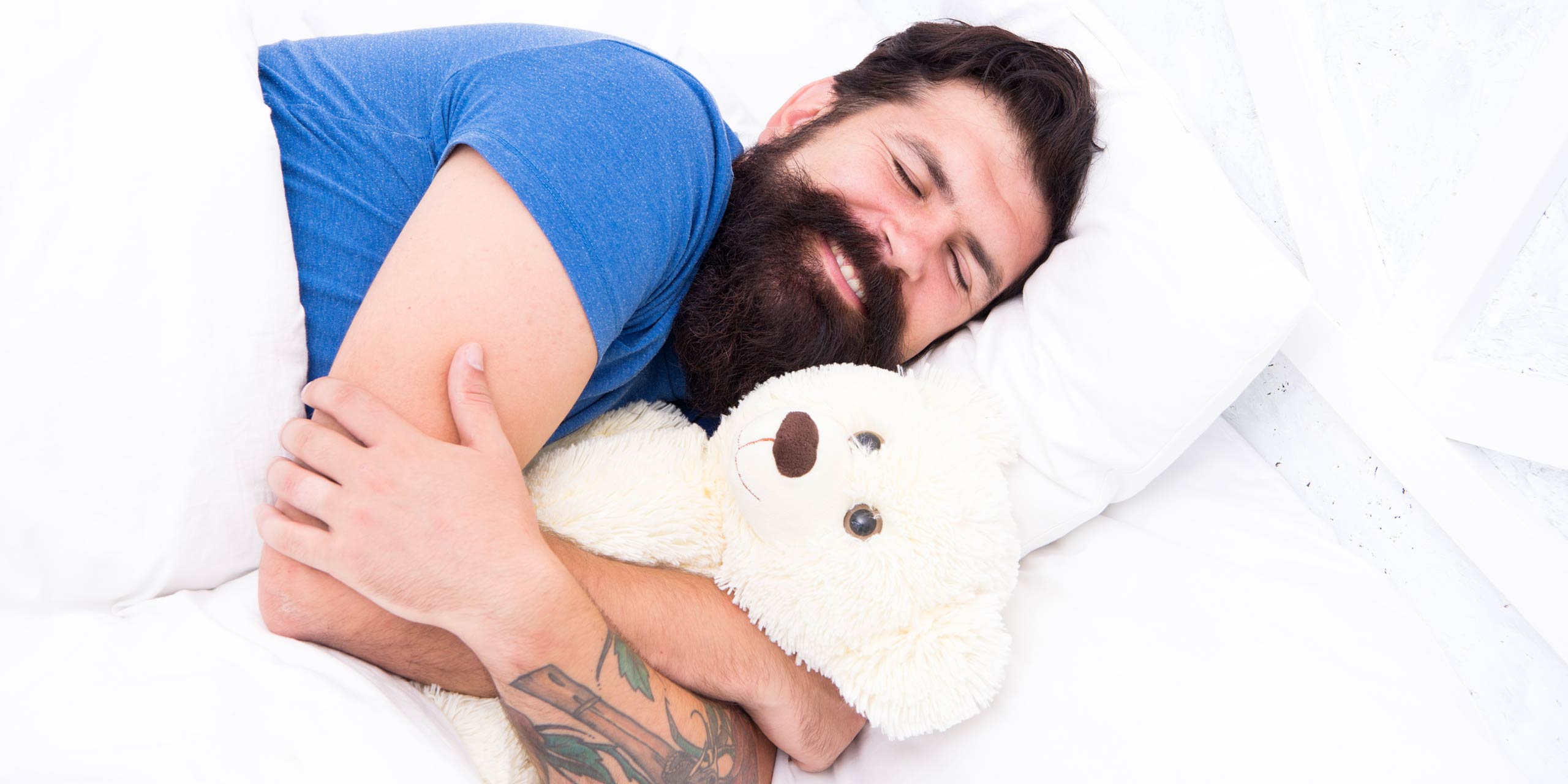 Mann mit Bart in blauem Shirt umarmt schlafend einen weißen Teddy.