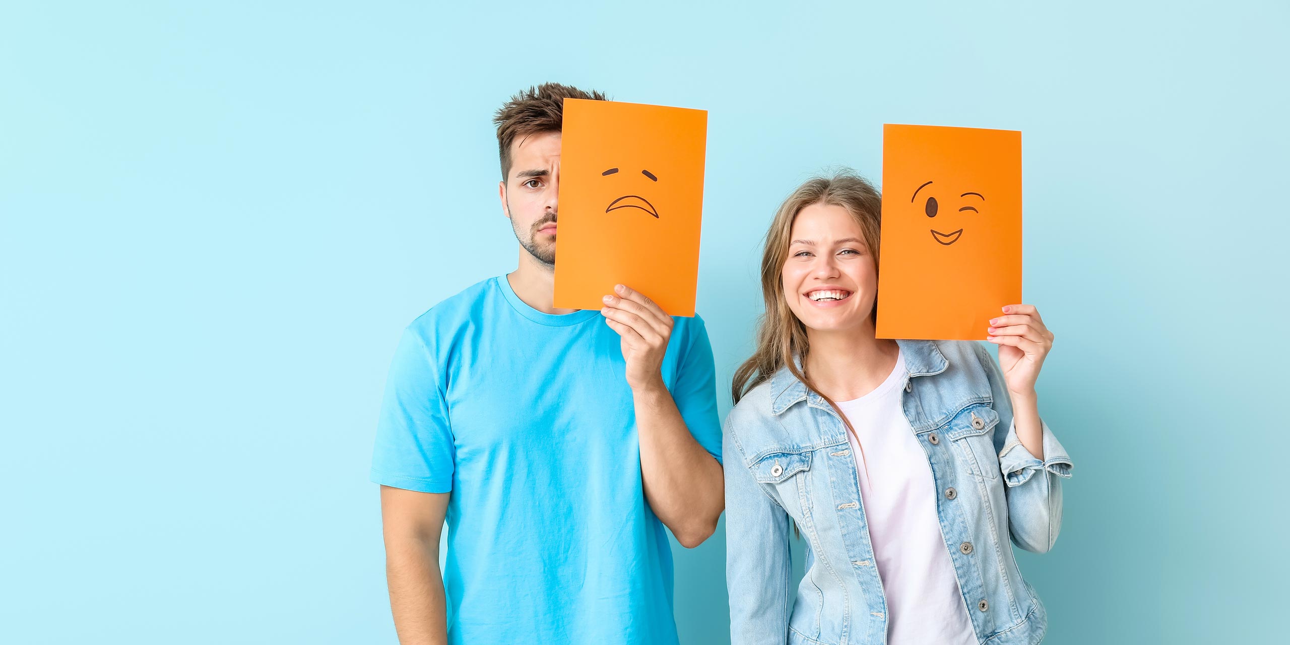 Junger Mann und junge Frau mit orangefarbenen Kartons, einer zeigt ein lächelndes, einer ein trauriges Gesicht.